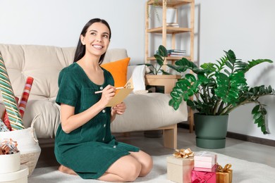 Photo of Happy woman writing message in greeting card on floor in living room