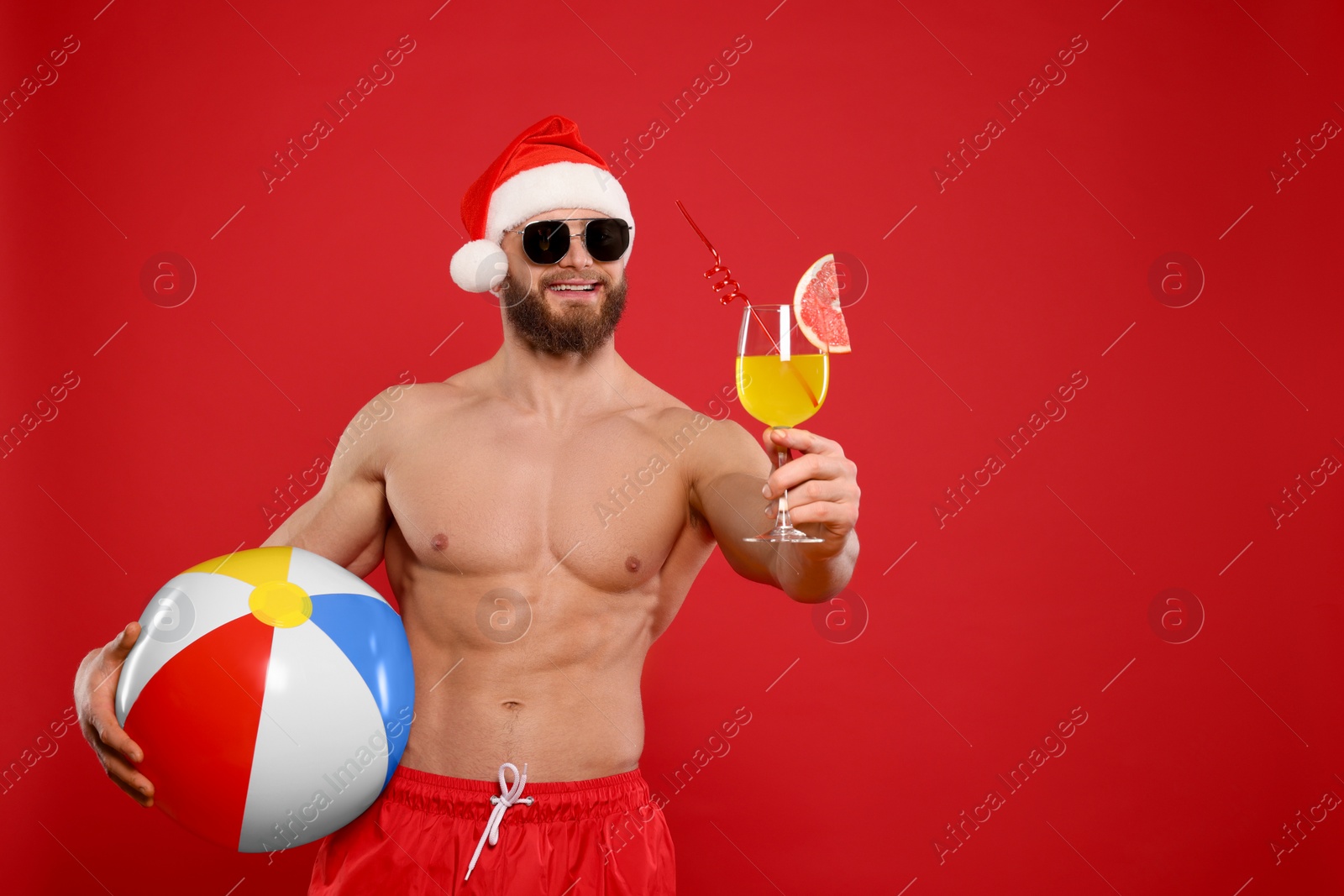 Photo of Muscular young man in Santa hat and sunglasses with ball and cocktail on red background, space for text