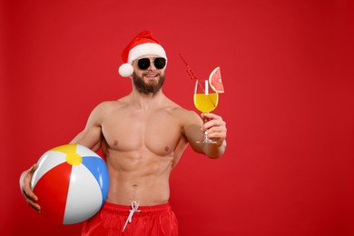 Photo of Muscular young man in Santa hat and sunglasses with ball and cocktail on red background, space for text
