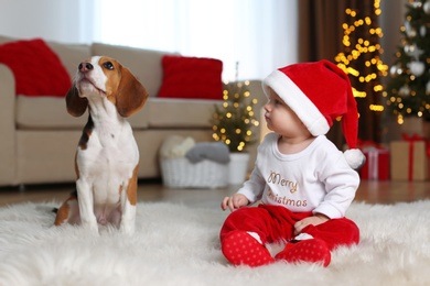 Photo of Baby in Santa hat and cute Beagle dog at home against blurred Christmas lights