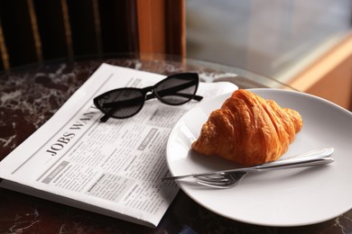 Tasty croissant, newspaper and sunglasses on black table