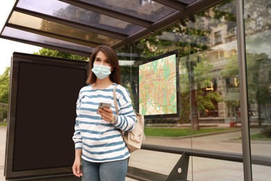 Image of Young woman in medical face mask waiting for public transport at bus stop