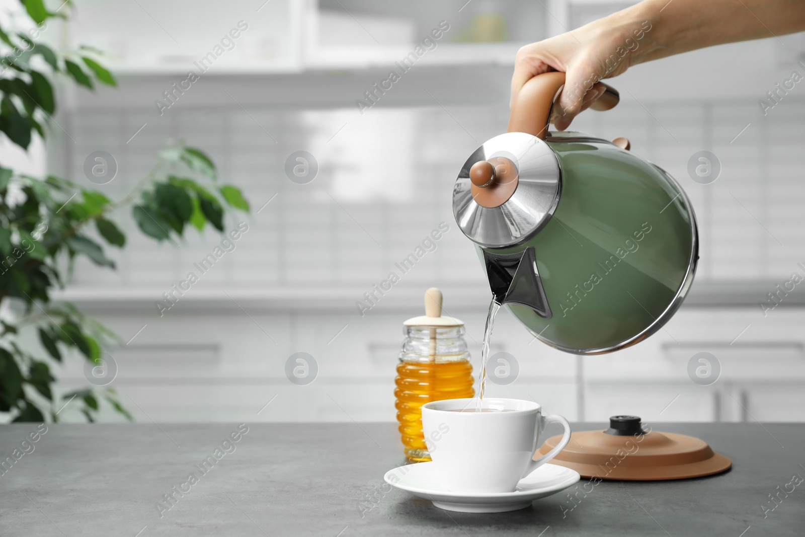 Photo of Woman pouring water from modern electric kettle into cup at grey table in kitchen, closeup. Space for text