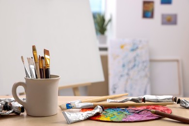 Photo of Art supplies on table near easel with canvas indoors. Space for text