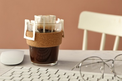 Glass cup with drip coffee bag, keyboard and glasses on white table, closeup