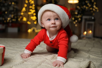 Cute little baby in Santa Claus suit on blanket against blurred festive lights. Christmas celebration