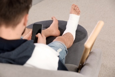 Young man with broken leg in cast using mobile phone while sitting on sofa at home