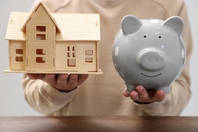 Photo of Man holding house model and piggy bank at wooden table, closeup. Saving money concept