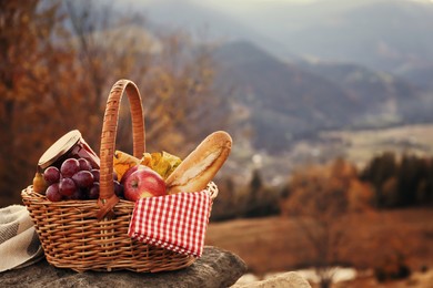 Image of Wicker picnic basket with different products on rock in mountains. Space for text
