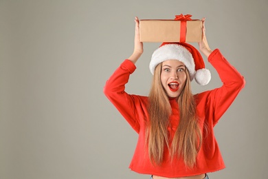 Young beautiful woman in Santa hat with gift box on grey background. Christmas celebration