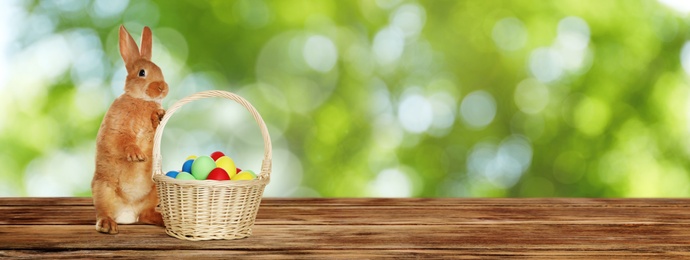 Adorable bunny and wicker basket with Easter eggs on wooden surface outdoors, space for text. Banner design