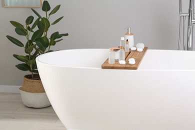 Photo of Wooden tray with cosmetic products and candles on tub in bathroom