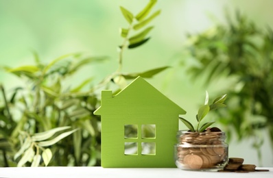 Model of house near jar with coins and plant on table against blurred white background. Space for text