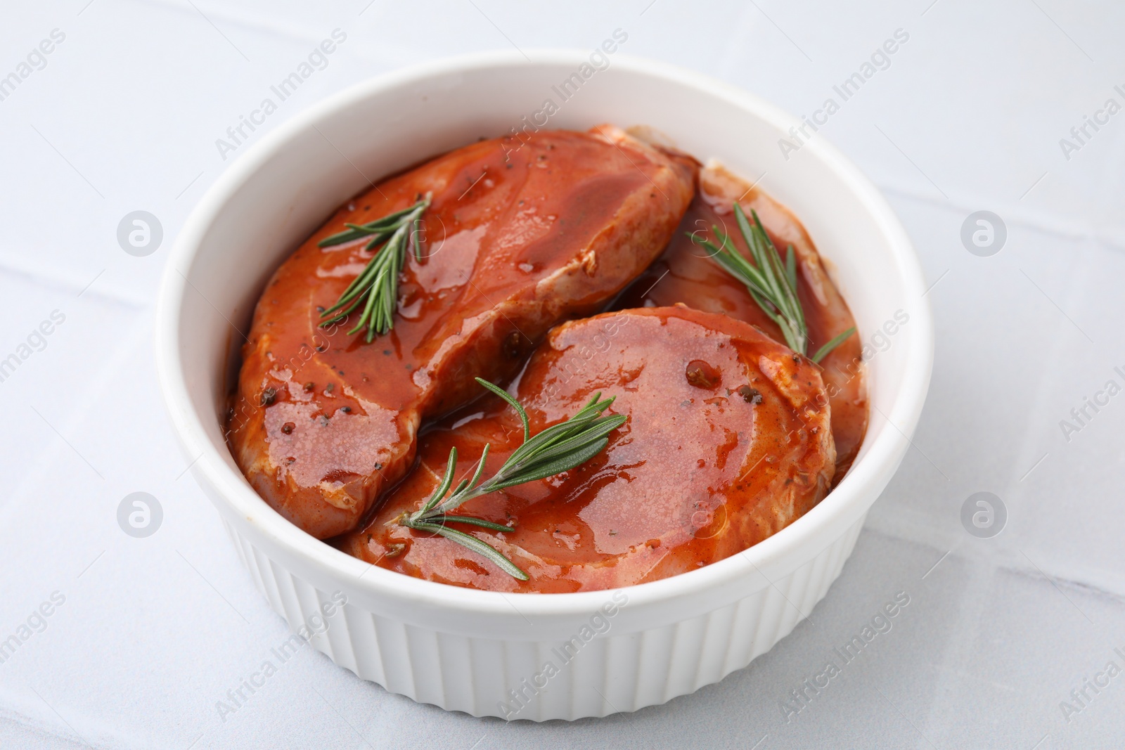 Photo of Raw marinated meat and rosemary in bowl on white tiled table, closeup