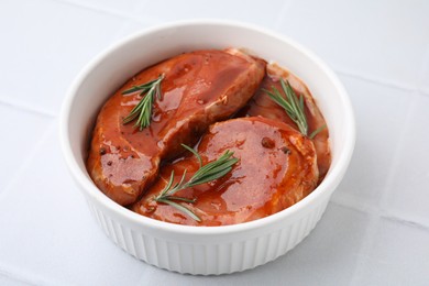Photo of Raw marinated meat and rosemary in bowl on white tiled table, closeup