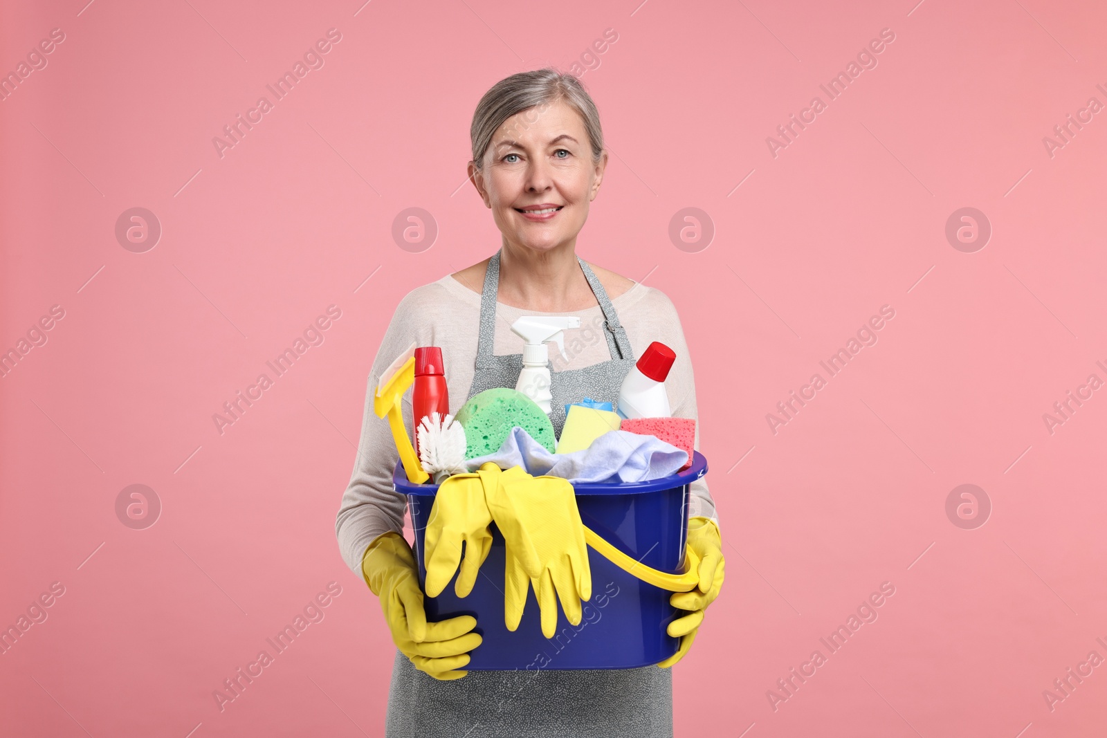 Photo of Happy housewife holding bucket with cleaning supplies on pink background