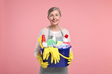 Photo of Happy housewife holding bucket with cleaning supplies on pink background