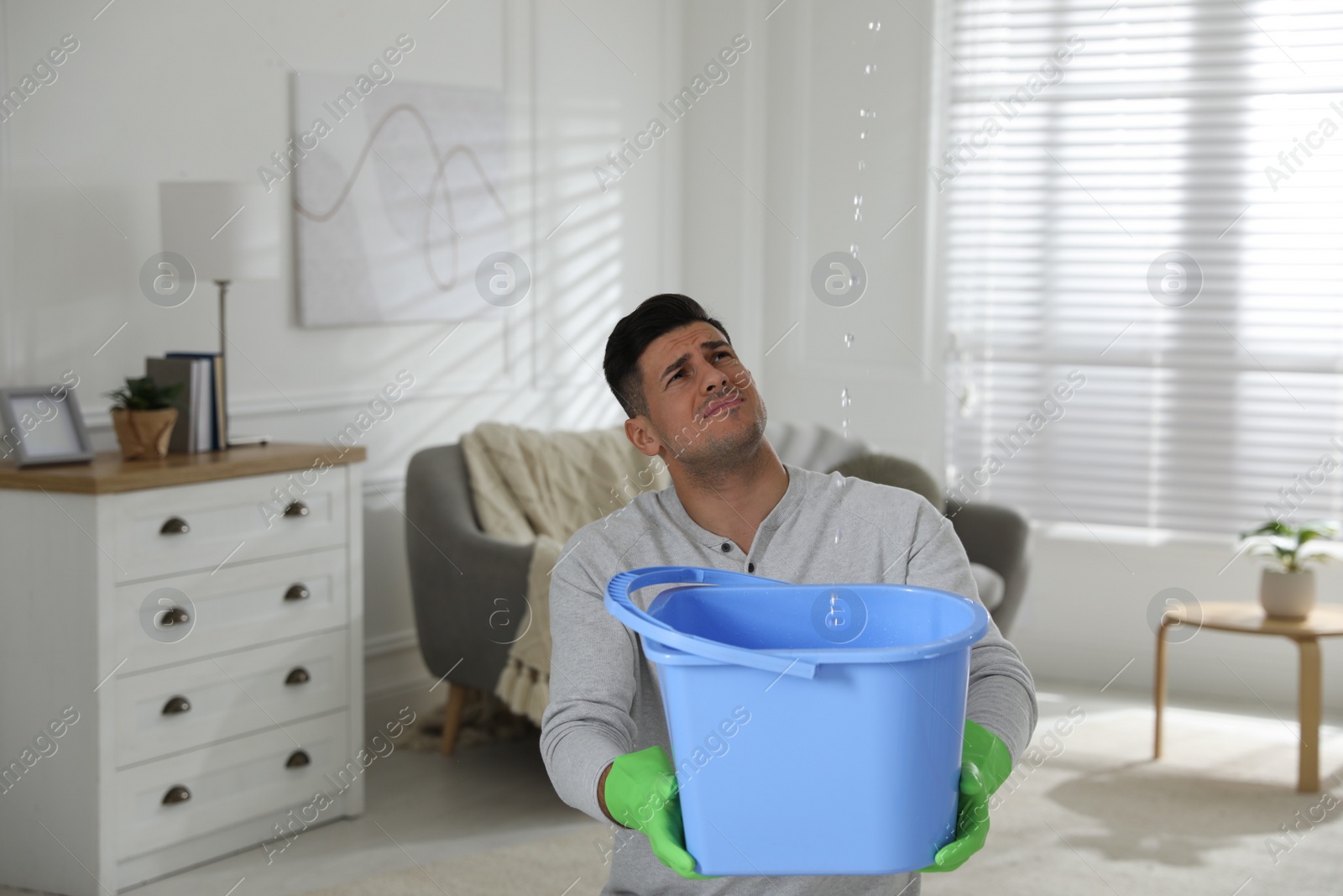 Photo of Emotional man collecting water leaking from ceiling in living room. Damaged roof