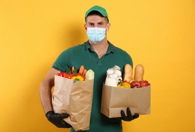 Courier in medical mask holding paper bags with food on yellow background. Delivery service during quarantine due to Covid-19 outbreak