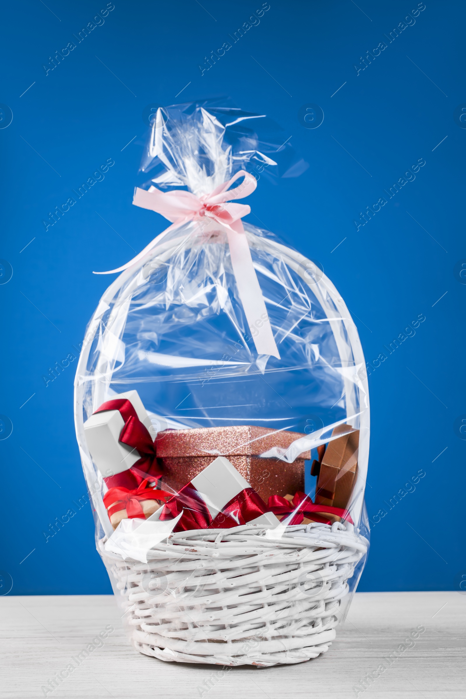 Photo of Wicker basket full of gift boxes on white wooden table against blue background