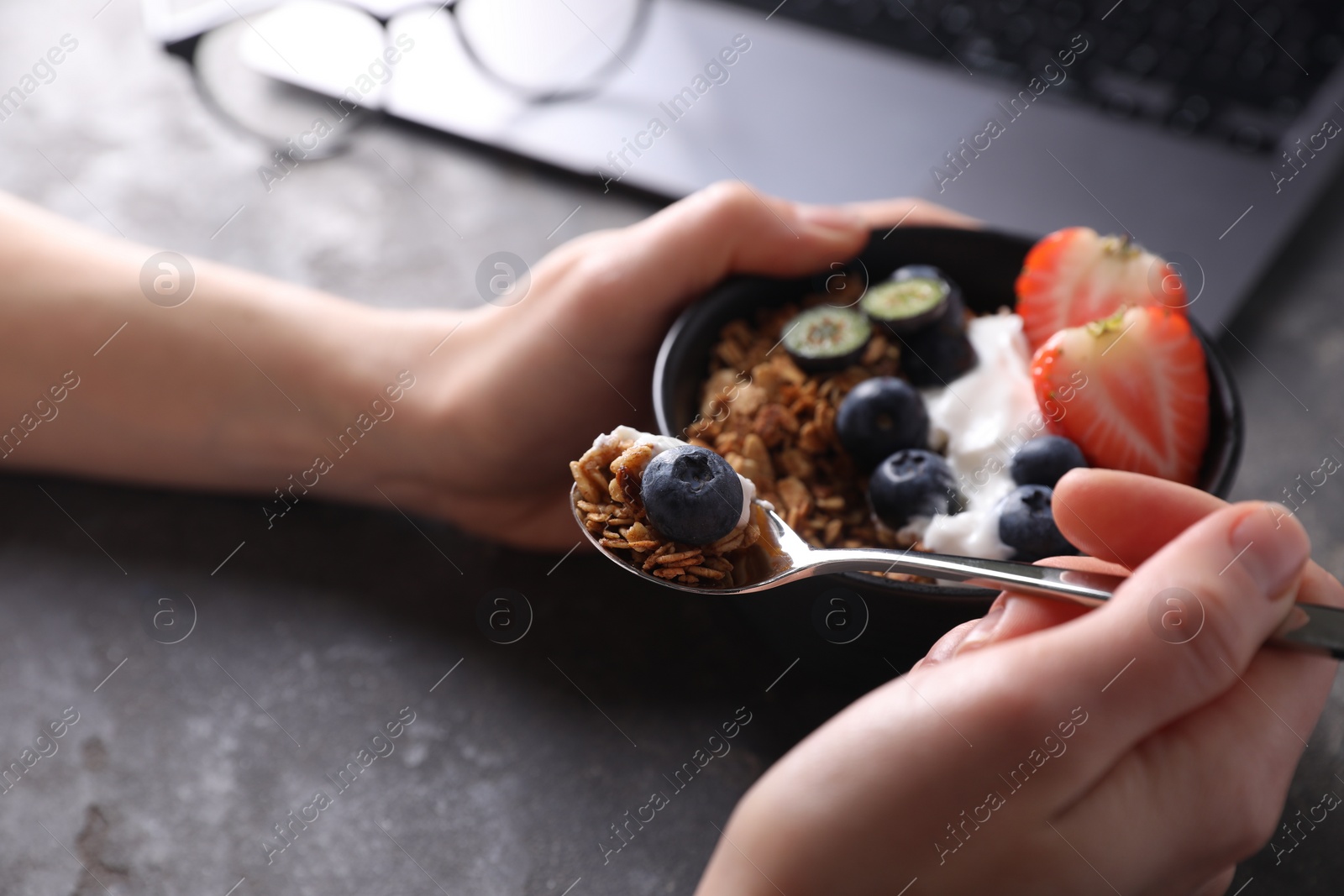 Photo of Woman eating tasty granola with yogurt and berries at workplace, closeup