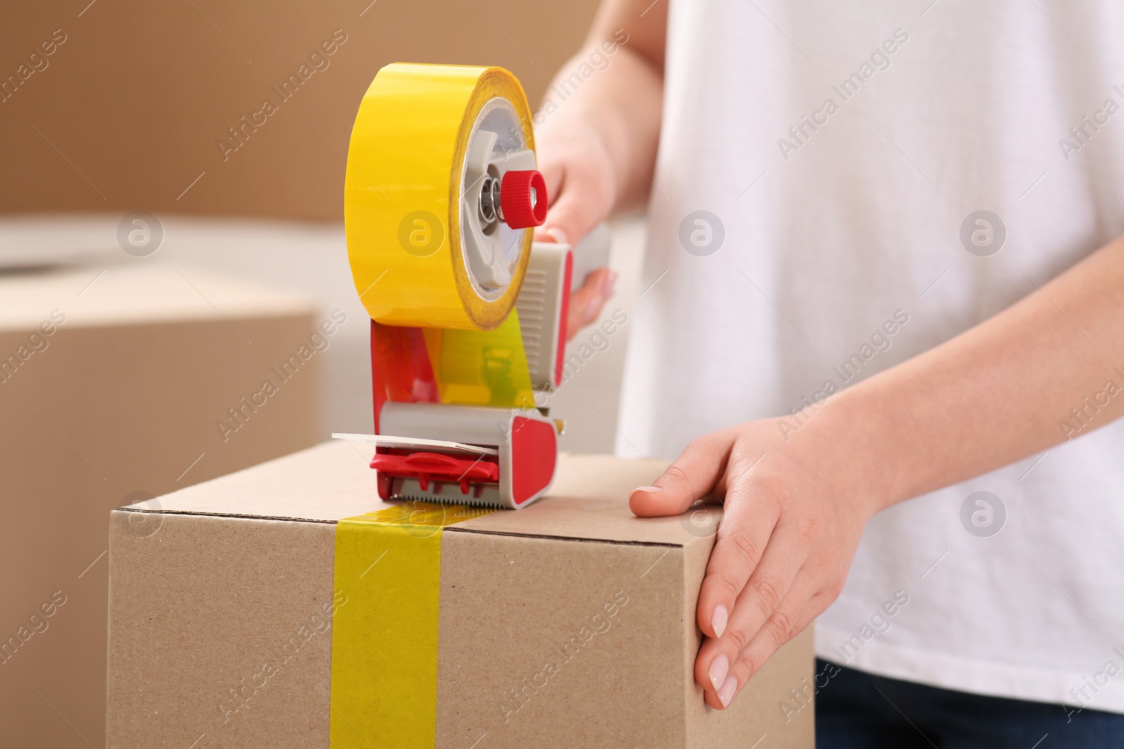 Photo of Woman applying adhesive tape on box with dispenser indoors, closeup