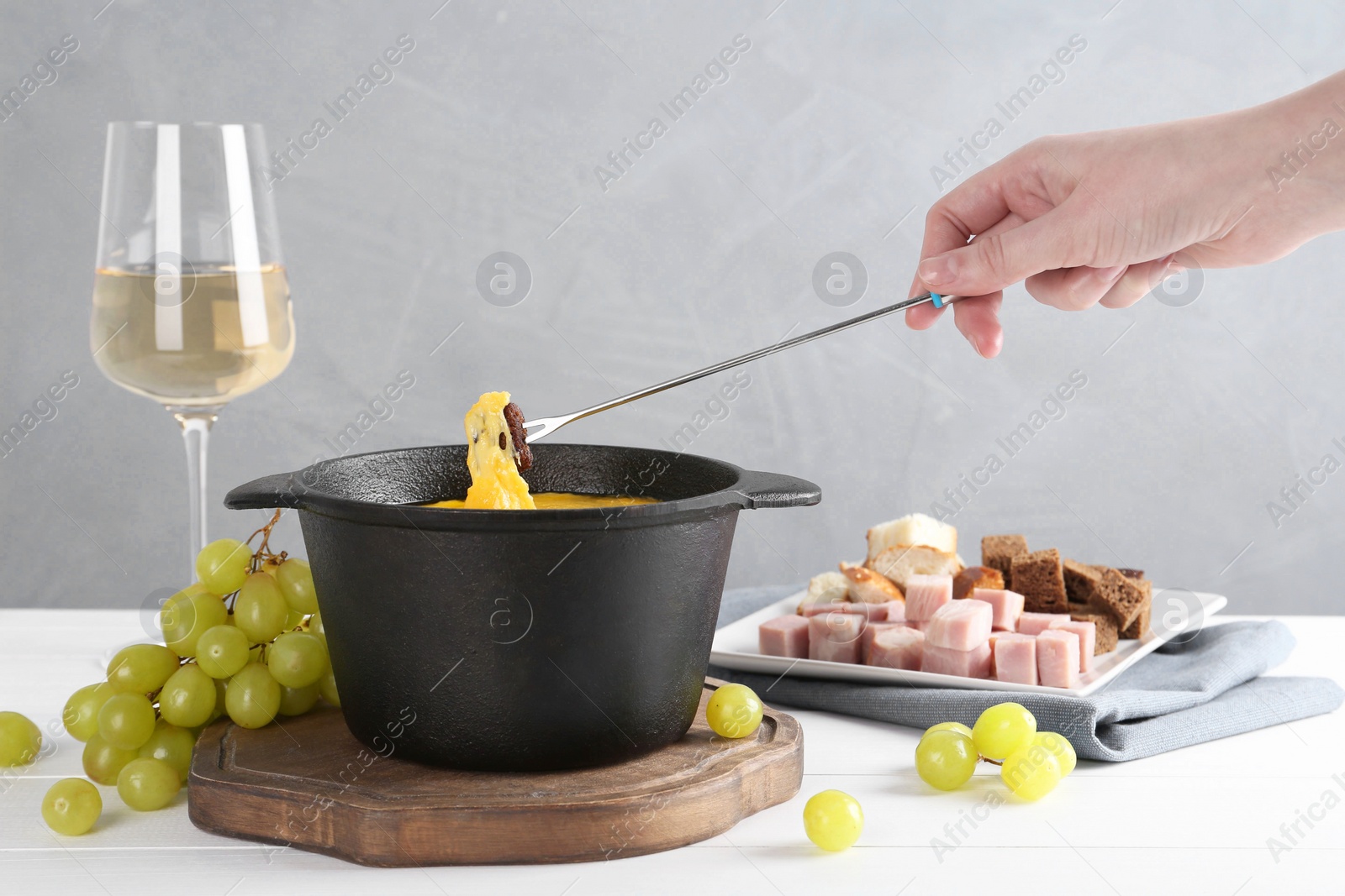 Photo of Woman dipping piece of bread into fondue pot with tasty melted cheese at white wooden table against gray background, closeup