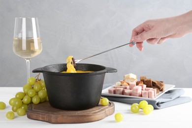 Woman dipping piece of bread into fondue pot with tasty melted cheese at white wooden table against gray background, closeup