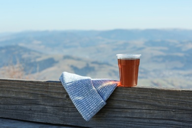 Photo of Plastic cup of natural hot tea and hat on wooden railing in mountains. Space for text