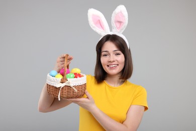 Photo of Easter celebration. Happy woman with bunny ears and wicker basket full of painted eggs on grey background