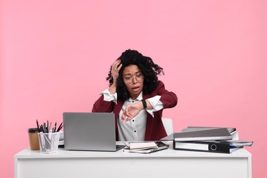 Stressful deadline. Scared woman checking time on wristwatch at white desk against pink background