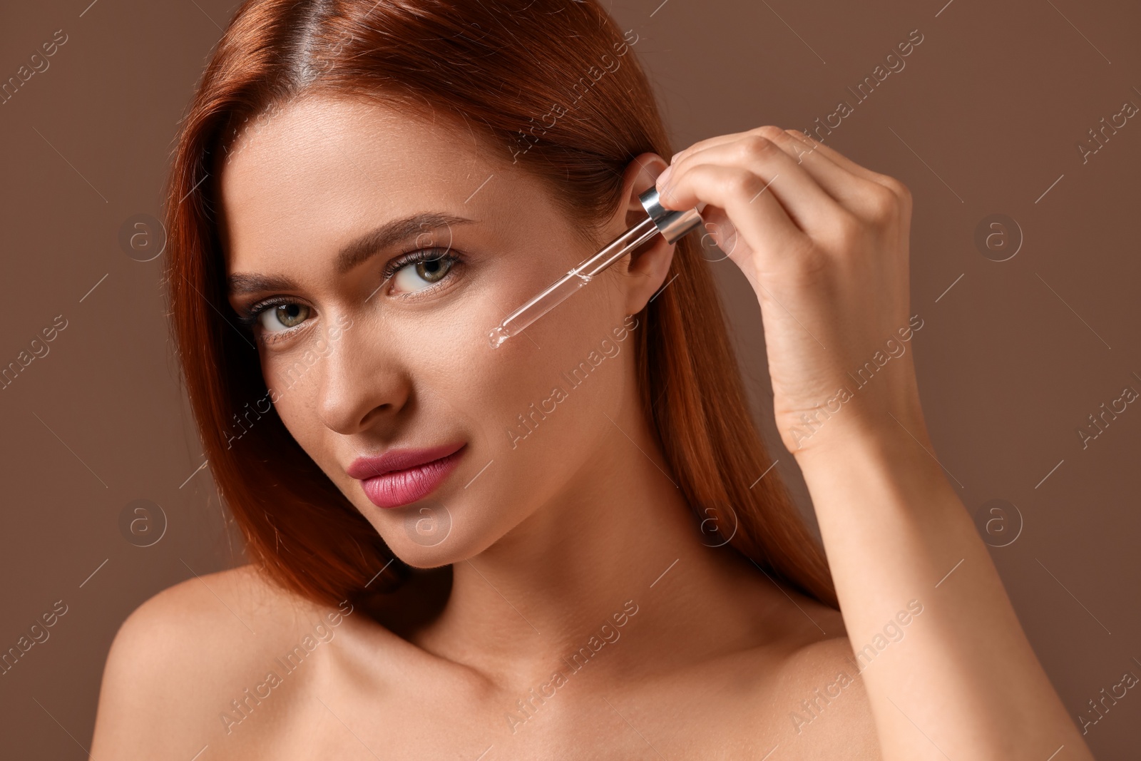 Photo of Beautiful young woman applying cosmetic serum onto her face on brown background