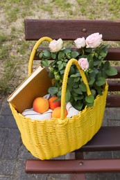 Yellow wicker bag with roses, book and peaches on bench outdoors