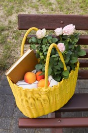 Photo of Yellow wicker bag with roses, book and peaches on bench outdoors