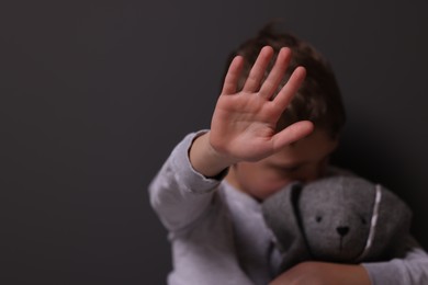 Child abuse. Boy with toy bunny doing stop gesture near gray wall, selective focus. Space for text