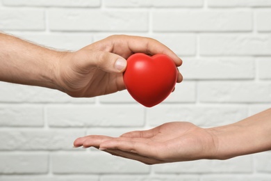 Man giving red heart to woman at white brick wall, closeup. Donation concept