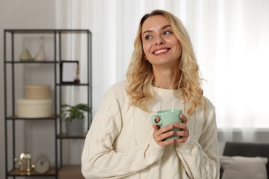 Photo of Happy woman in stylish warm sweater holding cup of drink at home