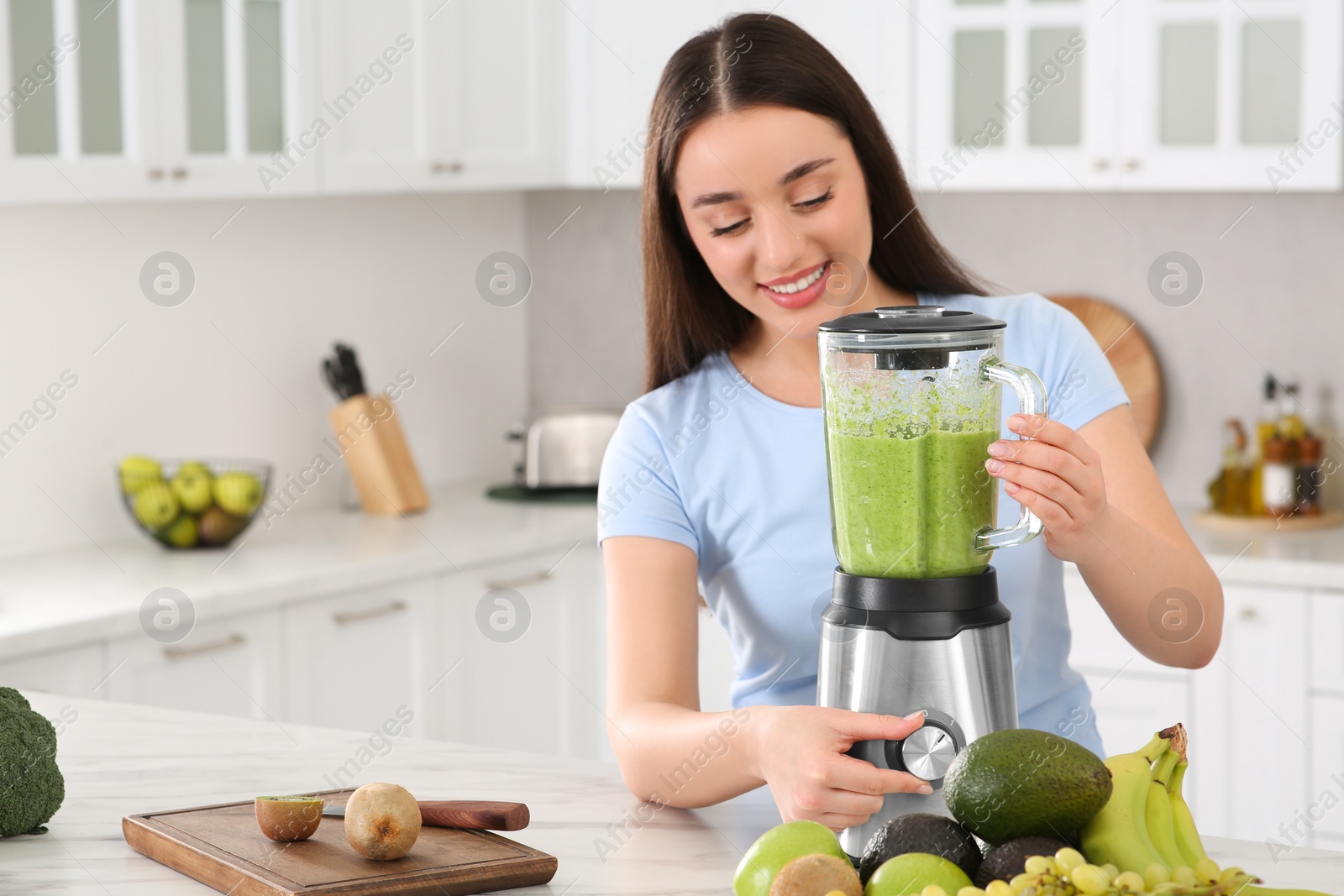 Photo of Beautiful young woman preparing tasty smoothie at white table in kitchen