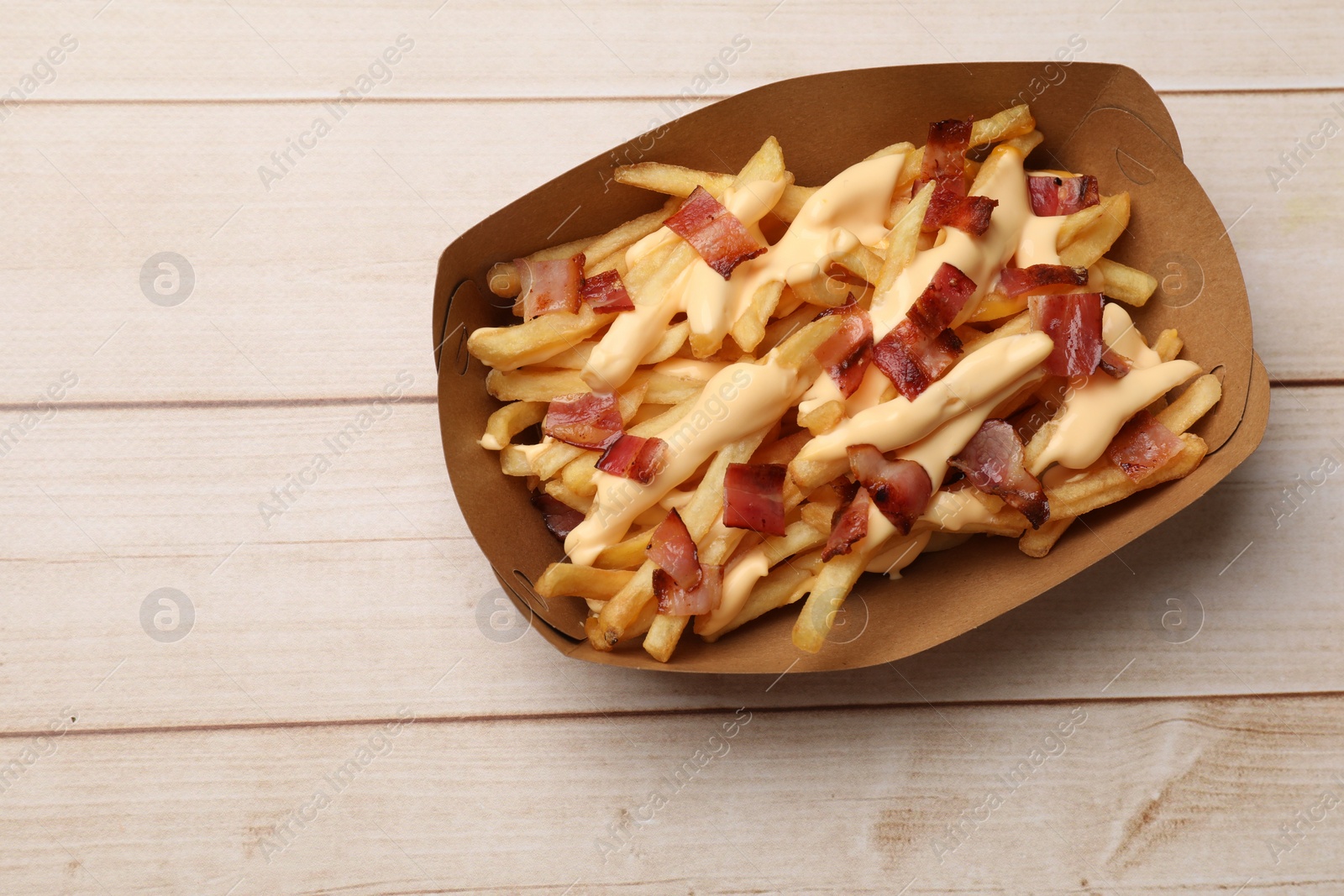 Photo of Tasty potato fries, cheese sauce and bacon in paper container on light wooden table, top view. Space for text