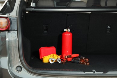 Photo of Red fire extinguisher, battery jumper cables, towing strap and first aid kit in trunk. Car safety