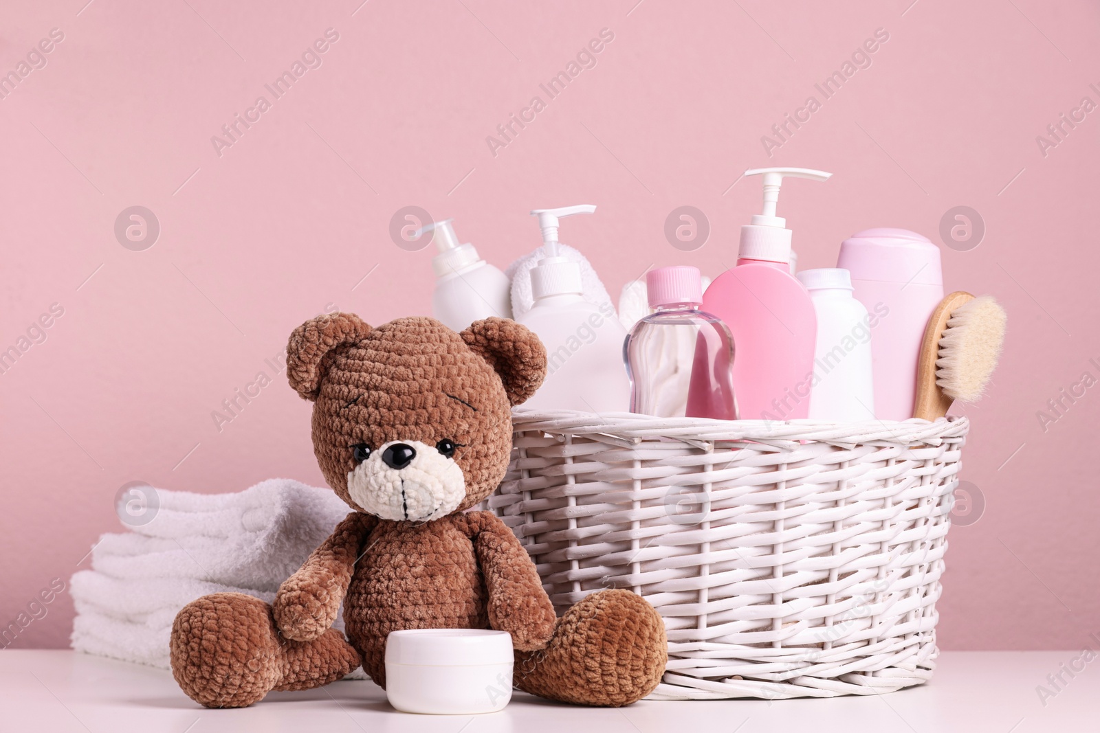 Photo of Baby cosmetic products in wicker basket, bath accessories and knitted toy bear on white table against pink background