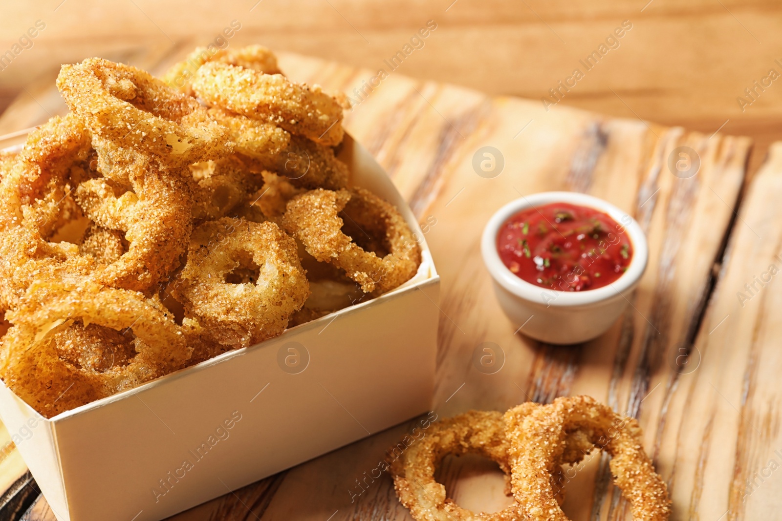 Photo of Cardboard box with crunchy fried onion rings and tomato sauce on wooden board