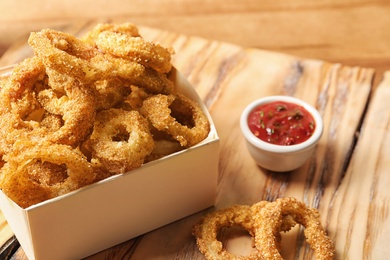 Photo of Cardboard box with crunchy fried onion rings and tomato sauce on wooden board