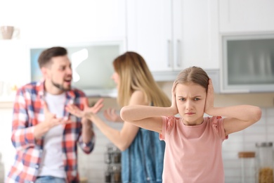 Photo of Little unhappy girl sitting at table while parents arguing on kitchen