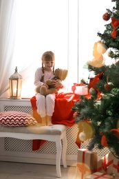 Cute little child with toy rabbit sitting on windowsill near Christmas tree at home