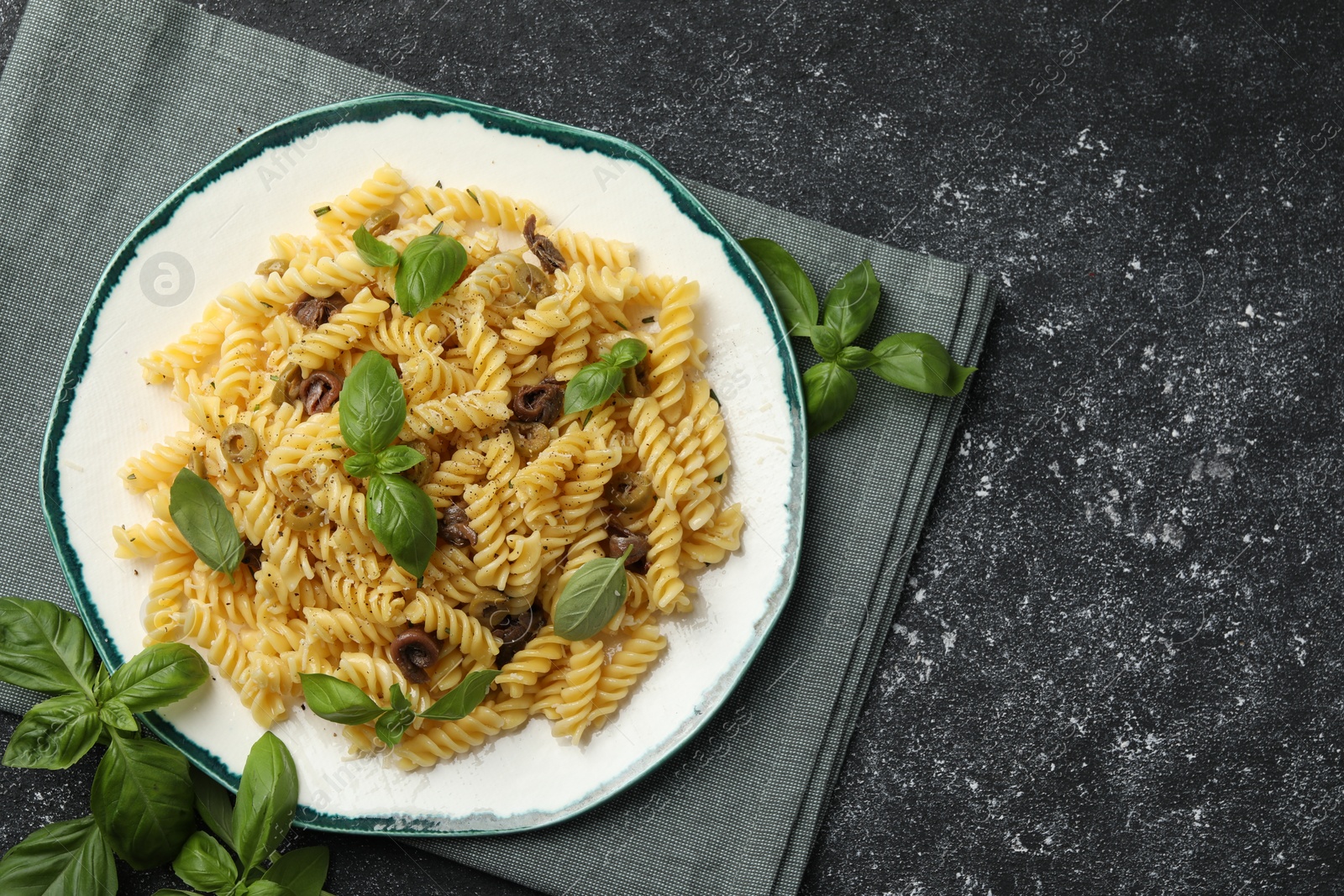 Photo of Plate of delicious pasta with anchovies and basil on black textured table, flat lay. Space for text