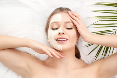 Photo of Beautiful woman with clay facial mask and tropical leaf on white fabric, above view