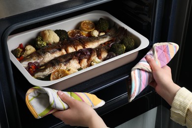Photo of Woman taking out baking tray with sea bass fish and vegetables from oven, closeup