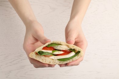 Photo of Woman holding delicious pita sandwich with mozzarella, tomatoes and basil at white wooden table, closeup