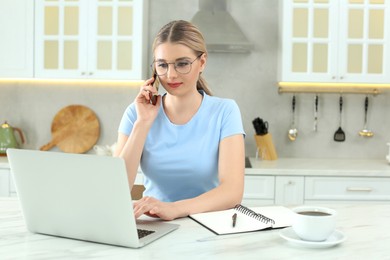 Home workplace. Woman talking on smartphone near laptop at marble desk in kitchen